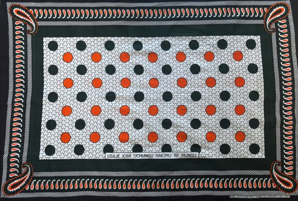 Kanga cloth from Tanzania showing the distinctive Swahili proverb printed along the fabric border, with bold geometric patterns in orange, blue and black.